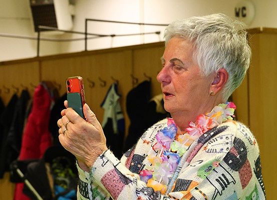 Eine ältere Frau mit weißen Haaren hält ein rotes Smartphone und schaut darauf. Sie trägt eine Blumenkette und einen Pullover. Hinter ihr befindet sich eine Holzwand mit hängenden Mänteln und einem weißen Schild.