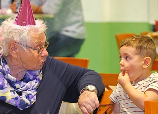Eine ältere Frau mit Partyhut und Brille spricht zu einem jungen Jungen, der seine Hand im Mund hat.