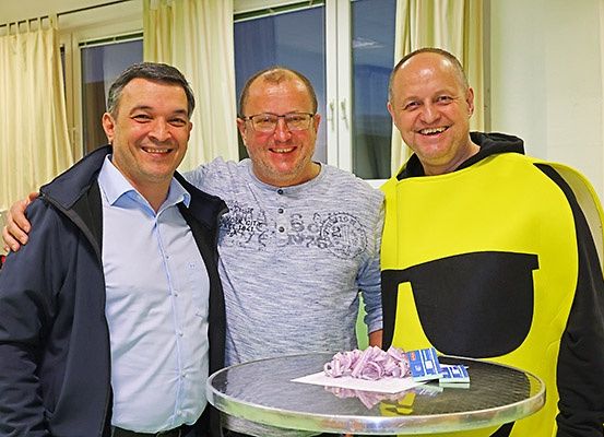 Drei Männer lächeln und posieren für ein Foto. Einer trägt eine blaue Jacke, ein anderer ein graues T-Shirt mit Brille und der dritte ein gelbes Gesichtskostüm. Ein Tisch vor ihnen hat einen Kuchen und Karten. Hinter ihnen befinden sich Fenster mit weißen Vorhängen.