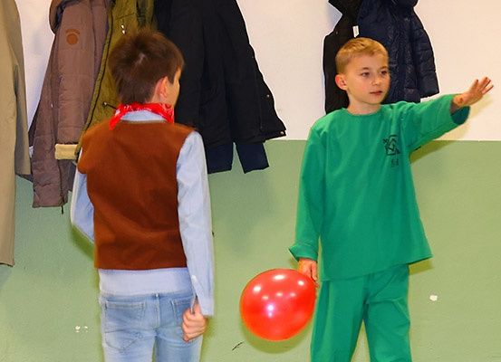 Zwei Jungen stehen in einem Raum, einer hält einen roten Luftballon. Der Junge in Grün schaut auf den Ballon.