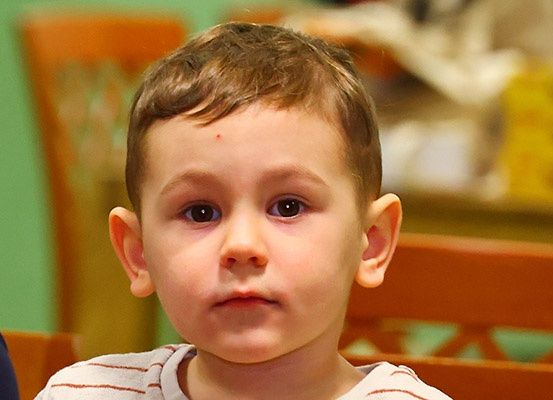 Ein junger Junge mit braunen Augen und einem kurzen Haarschnitt schaut direkt in die Kamera. Er trägt ein weißes Hemd mit roten Streifen.