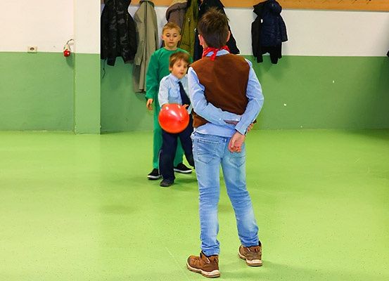 Drei Kinder spielen mit einem roten Ball in einem Raum mit grünem Boden. Hinter ihnen hängen Kleidungsstücke an der Wand.