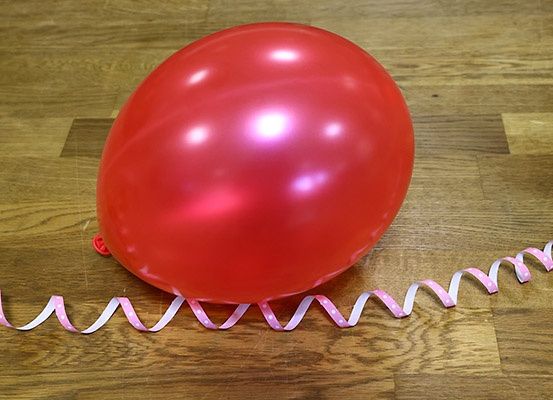 Ein roter Ballon mit einer rosa Schleife auf einem Holzboden, der Licht reflektiert. Der Ballon ist vollständig aufgeblasen.