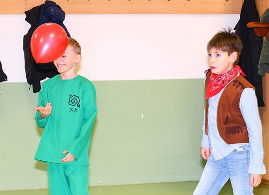 Zwei junge Jungen in Kostümen, einer hält einen roten Ballon, stehen in einem Raum mit grünem Boden.