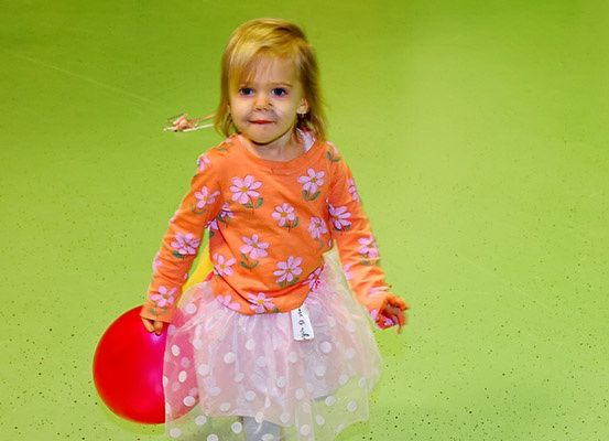 Ein kleines Mädchen mit blonden Haaren steht auf einem grünen Boden, hält einen roten Ballon und trägt eine Tulpe. Sie scheint zu lächeln.