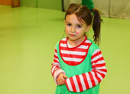 Ein kleines Mädchen mit Zöpfen und rosa Farbe auf den Wangen trägt ein grün-weiß gestreiftes Shirt und Kleid. Sie steht in einem Raum mit grünem Boden.
