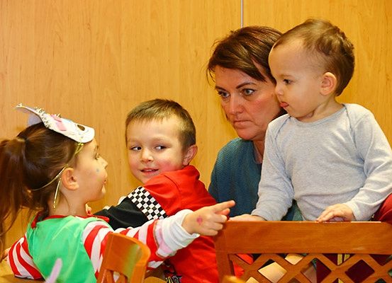 Eine Frau und zwei kleine Kinder sitzen an einem Tisch, wobei ein Kind auf etwas zeigt und das andere etwas ansieht. Die Frau lächelt.