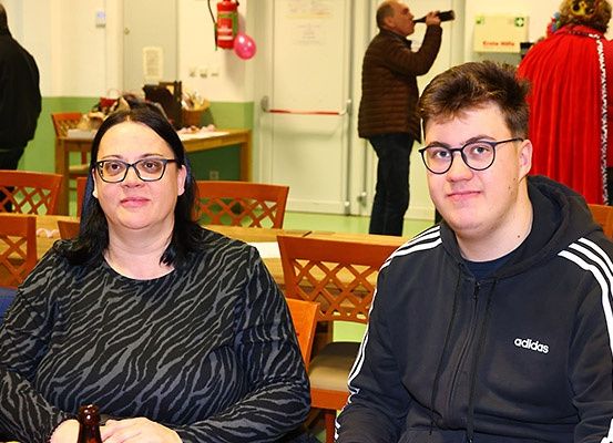 Ein Mann und eine Frau sitzen an einem Tisch in einem Raum mit Stühlen, einem Tisch und Personen im Hintergrund.
