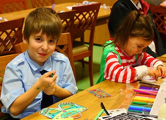 Zwei Kinder sitzen an einem Tisch und beschäftigen sich mit dem Ausmalen. Der Junge hat farbige Designs an den Armen und hält einen Marker. Das Mädchen trägt ein gestreiftes Shirt und hält ein Malbuch.