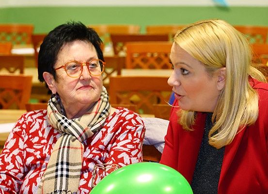 Zwei Frauen sitzen in einem Klassenzimmer mit leeren Stühlen, eine mit Brille und Schal, die andere hält einen grünen Luftballon.