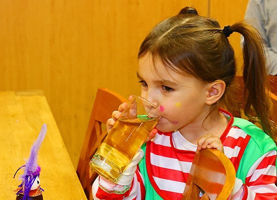 Ein kleines Mädchen trinkt aus einem Glas mit gelber Flüssigkeit und trägt ein rot-weiß gestreiftes Shirt. Sie sitzt an einem Holztisch mit einem Holzstuhl hinter ihr.