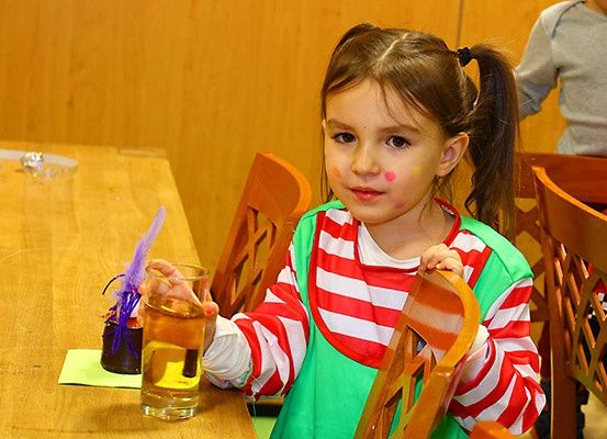 Ein kleines Mädchen mit rot-weiß gestreiften Ärmeln und einer grünen Schürze hält ein Glas mit Flüssigkeit. Dahinter ist ein Tisch mit einem gelben Papier und einem kleinen schwarzen Gegenstand zu sehen.