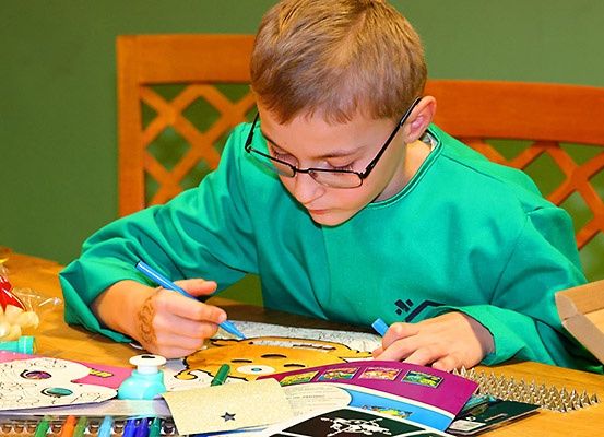 Ein junger Junge mit Brille färbt ein Bild mit verschiedenen Farben ein. Er sitzt an einem Tisch mit einem Malbuch, Markern und anderen Malutensilien. Hinter ihm steht ein Holzstuhl und eine grüne Wand.