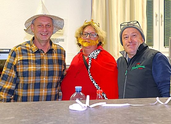 Drei Männer stehen zusammen in einem Raum. Der Mann auf der linken Seite trägt ein kariertes Hemd und einen Hut. Der Mann in der Mitte trägt einen roten Umhang mit einem weißen Leopardenmuster und eine Krone. Der Mann auf der rechten Seite trägt eine graue Mütze und eine Jacke mit dem Wort 'Nockspitze' darauf. Vor ihnen stehen zwei Flaschen und ein Band. Hinter ihnen befindet sich eine Wand mit einer Tafel.