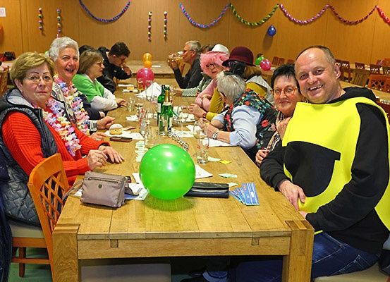 Eine Gruppe von Menschen sitzt an einem langen Tisch in einem Esszimmer. Sie tragen festliche Kleidung und einige tragen Hüte. Sie lächeln und posieren für ein Foto. Der Tisch ist mit verschiedenen Gegenständen gefüllt, darunter Ballons, Gläser und Teller. Der Raum ist mit Ballons und Girlanden dekoriert.