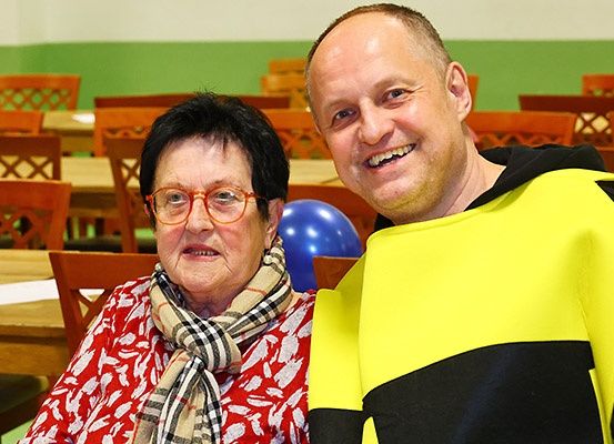 Eine Frau mit Brille und einem karierten Schal und ein Mann in einem gelben und schwarzen Outfit lächeln in einem Essbereich.