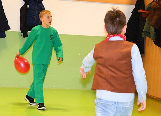 Zwei junge Jungen spielen mit einem roten Ballon in einem grünen Raum. Ein Junge trägt grüne Kleidung, der andere trägt eine braune Weste und ein rotes Halstuch.