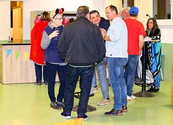 Eine Gruppe von Menschen, einige in Kostümen gekleidet, stehen und unterhalten sich in einem Raum mit Theke und Luftballons.