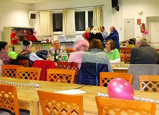Mehrere Erwachsene sind in einem Essbereich versammelt, einige tragen Partyhüte. Der Raum ist mit Luftballons und Girlanden dekoriert. Tische und Stühle sind in Reihen angeordnet. Eine Person in einem roten Shirt steht hinter der Theke.