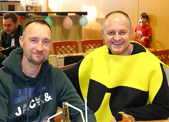 Zwei Männer in einem Restaurant, einer trägt eine grüne Kapuzenjacke und der andere ein gelbes und schwarzes Kostüm, lächeln in die Kamera. Hinter ihnen steht ein Kind am Tisch.