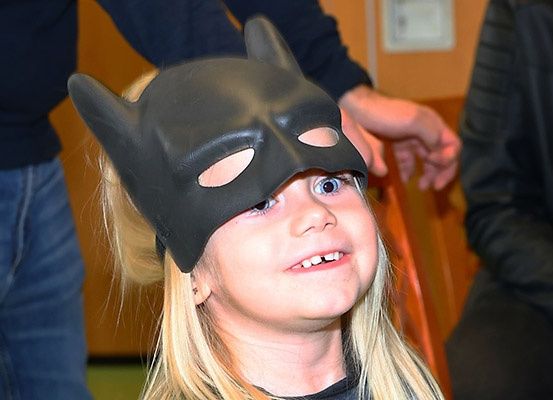 Ein kleines Mädchen mit blonden Haaren, das eine schwarze Batman-Maske trägt und lächelt, mit einer Person hinter ihr.