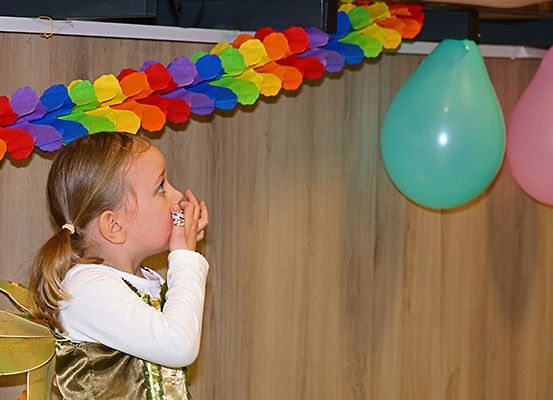 Ein kleines Mädchen in einem Kostüm bläst einen grünen Ballon auf in einem Raum mit einer bunten Girlande und zwei hängenden Ballons.