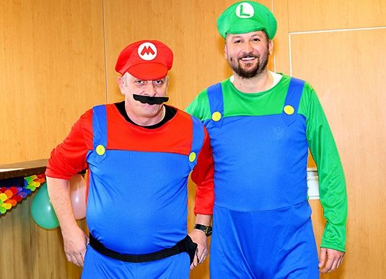 Zwei Männer in Mario- und Luigi-Kostümen stehen nebeneinander und lächeln. Hinter ihnen befindet sich eine Holzwand mit Luftballons.