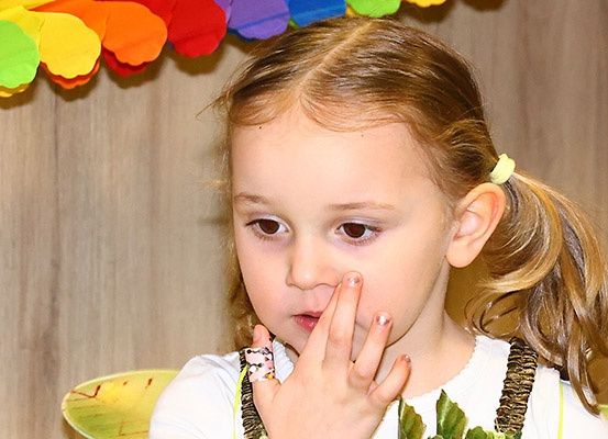 Ein junges Mädchen mit blondem Haar trägt ein weißes T-Shirt und hat den Finger auf der Nase, wahrscheinlich auf einer Party. Der Hintergrund zeigt eine bunte Papiergirlande.