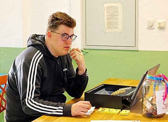 Ein Mann mit Brille und Kapuzenpullover sitzt an einem Schreibtisch, hält ein Stück Papier in der Hand und schaut auf eine Schachtel Scrabble-Steine. Ein Laptop und ein Glas mit einer rosa Schleife stehen ebenfalls auf dem Schreibtisch.