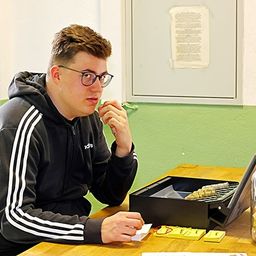 Ein Mann mit Brille und Kapuzenpullover sitzt an einem Schreibtisch, hält ein Stück Papier in der Hand und schaut auf eine Schachtel Scrabble-Steine. Ein Laptop und ein Glas mit einer rosa Schleife stehen ebenfalls auf dem Schreibtisch.