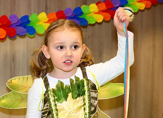 Ein kleines Mädchen in einem Schmetterlingskostüm, das ein Band hält, steht vor einem bunten Kranz.