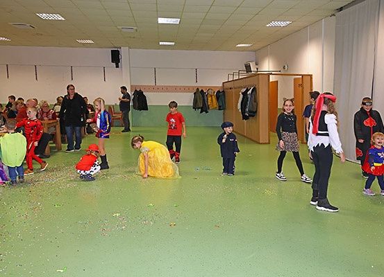 Im Inneren eines Raumes mit grünem Boden befindet sich eine Gruppe von Kindern in Kostümen, einige stehend, andere kriechend. Einige Erwachsene sind ebenfalls anwesend, und Jacken hängen an der Wand.
