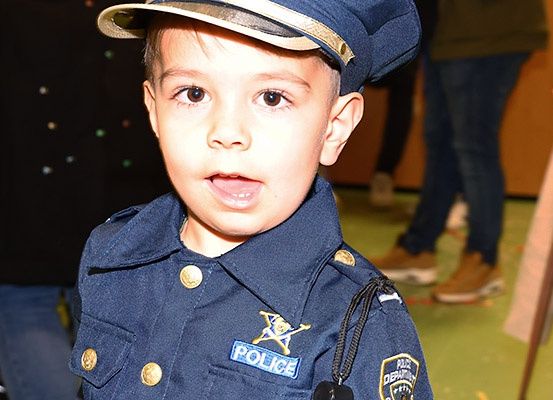 Ein junger Junge in einer Polizeiuniform, mit leicht geöffnetem Mund, umgeben von verschwommenen Figuren von Menschen im Hintergrund.