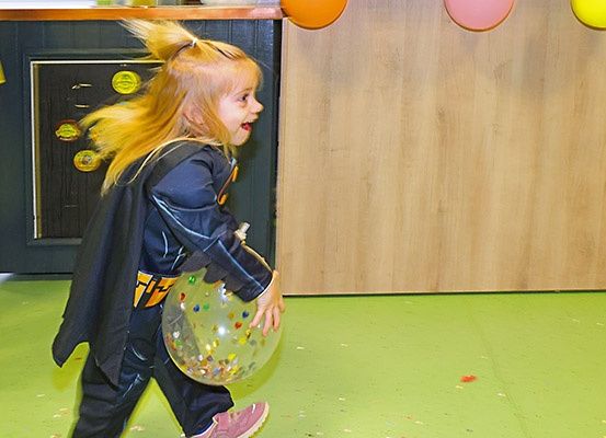 Ein kleines Mädchen in einem Superheldenkostüm läuft aufgeregt und hält einen mit bunten Murmeln gefüllten Ballon. Sie befindet sich in einem Raum mit grünem Boden und an der Decke hängenden Luftballons.