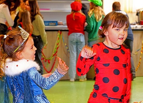 Zwei kleine Mädchen in Kostümen, eines als Prinzessin und das andere als Marienkäfer, tanzen in einem Raum mit anderen Leuten in Kostümen.