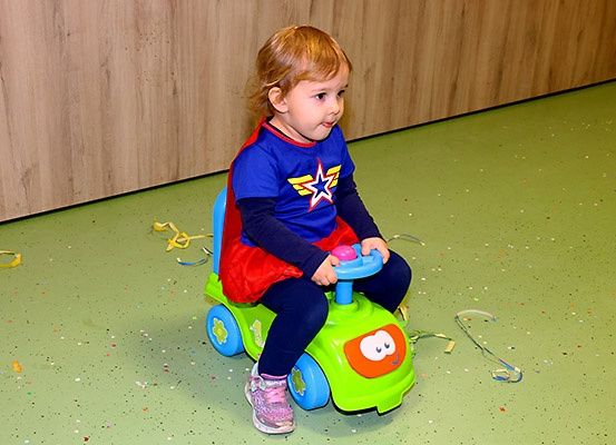 Ein kleines Mädchen in einem Superheldenkostüm sitzt auf einem Spielzeugauto und lächelt in einem Raum mit grünem Boden und Holzwänden.
