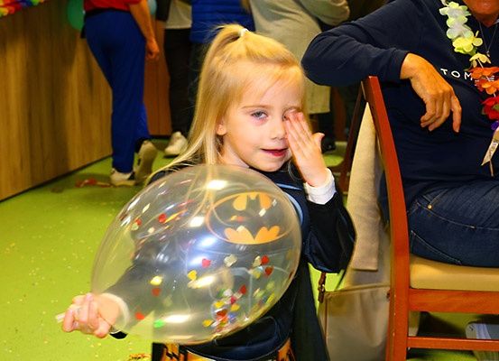 Ein kleines Mädchen, das als Batman verkleidet ist, hält einen bunten Ballon in Innenräumen, umgeben von anderen, einschließlich eines Erwachsenen mit einer Lei.