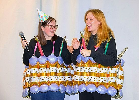 Zwei Frauen in Ballonkostümen, eine mit Mikrofon und die andere mit Kerzen, stehen nebeneinander in einem Raum.