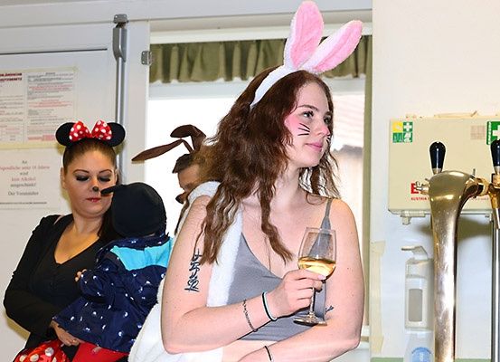 Eine Frau mit Hasenohren hält ein Glas Wein. Hinter ihr hält eine Frau mit Minnie-Mouse-Kopfband ein Kind. Ein weiteres Kind mit Hundekopfbedeckung steht neben ihnen.