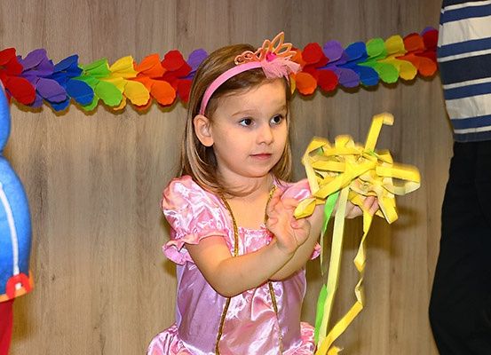Ein kleines Mädchen in einem rosa Kleid und Stirnband hält gelbe Streamer in einem Raum, der mit mehrfarbigen Papierblumen dekoriert ist. Eine Person in einem gestreiften Hemd und eine andere in roten Hosen sind ebenfalls anwesend.