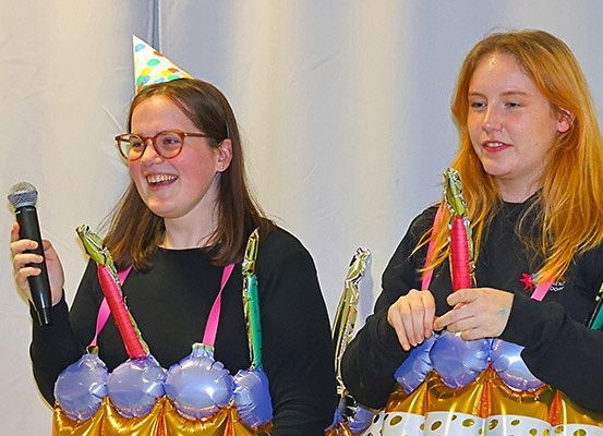 Zwei junge Frauen in Kostümen, eine hält ein Mikrofon und beide tragen bunte Ballonkostüme und Partyhüte. Sie scheinen auf einer Bühne aufzutreten.