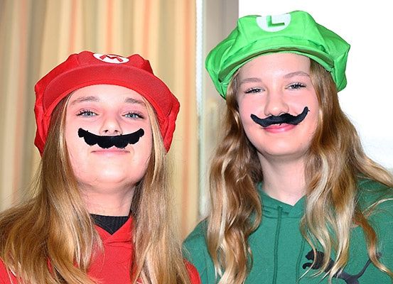 Zwei Mädchen in roten und grünen Kostümen mit gemalten Schnurrbärten auf den Gesichtern, die nebeneinander stehen.