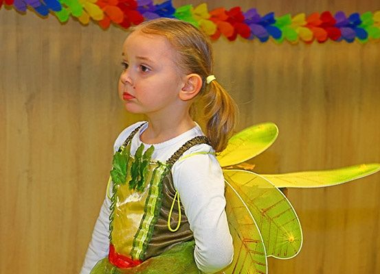 Ein junges Mädchen in einem grünen Feenkostüm mit Flügeln steht und schaut nach vorne. Hinter ihr befindet sich eine Holzwand mit einer bunten Girlande aus Papierblumen.