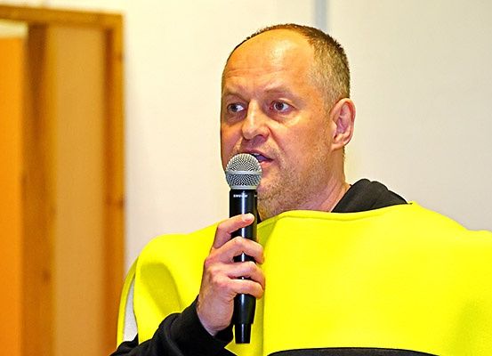 Ein Mann spricht in ein Mikrofon, trägt eine gelbe Warnweste und steht in einem Raum mit einer weißen Wand und einer Holztür.