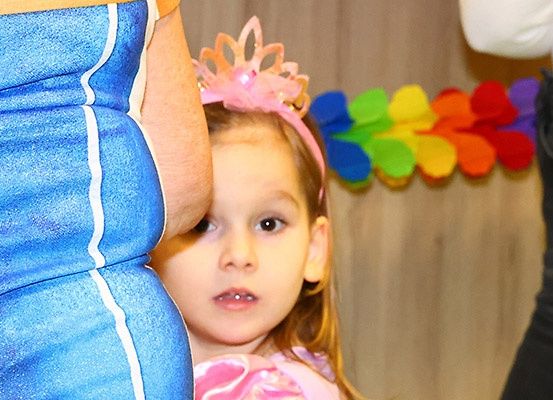 Ein kleines Mädchen mit einer rosa Prinzessinnen-Haarschleife steht neben einer Frau in einem blauen Kleid, mit einem bunten Kranz im Hintergrund.
