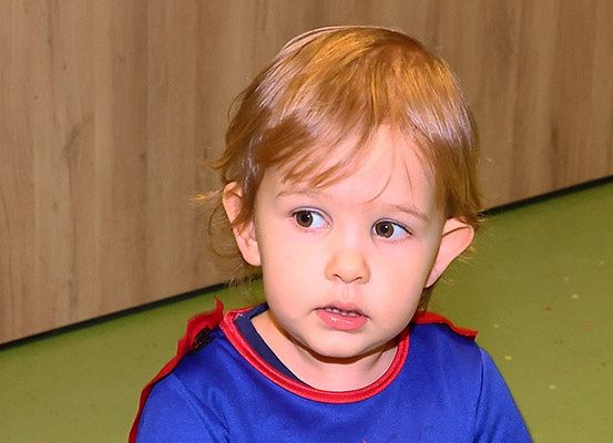 Ein junges Kind mit blondem Haar trägt ein blaues T-Shirt mit roten Einsätzen und sitzt auf einem grünen Boden. Es schaut mit leicht nach unten gezogenem Mund nach vorne. Hinter ihm befindet sich eine Holzwand.