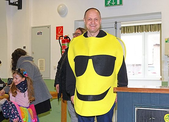 Ein Mann in einem gelben Smiley-Gesichtskostüm steht in einem Raum mit einem Feuerlöscher an der Wand. Hinter ihm sind eine Frau und ein junges Mädchen anwesend.