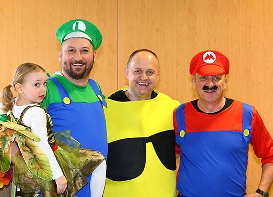 Drei Männer und ein Mädchen in Kostümen stehen zusammen. Die Männer sind als Mario und Luigi verkleidet, während das Mädchen ein Schmetterlingskostüm trägt. Sie lächeln für ein Foto.