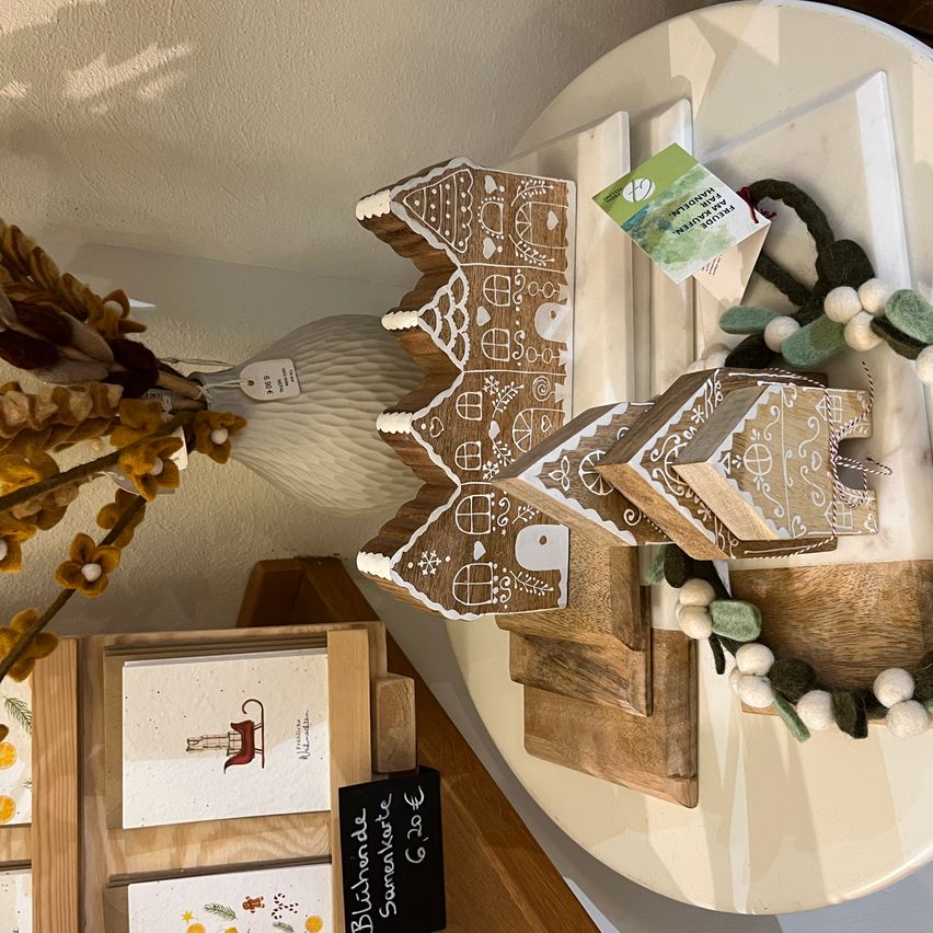 Ein Lebkuchenhaus steht prominent auf einem Tisch, umgeben von dekorativen Elementen. Ein weißer Vase mit einem Blumenarrangement steht in der Nähe. Es gibt Preisschilder und Karten zur Schau.