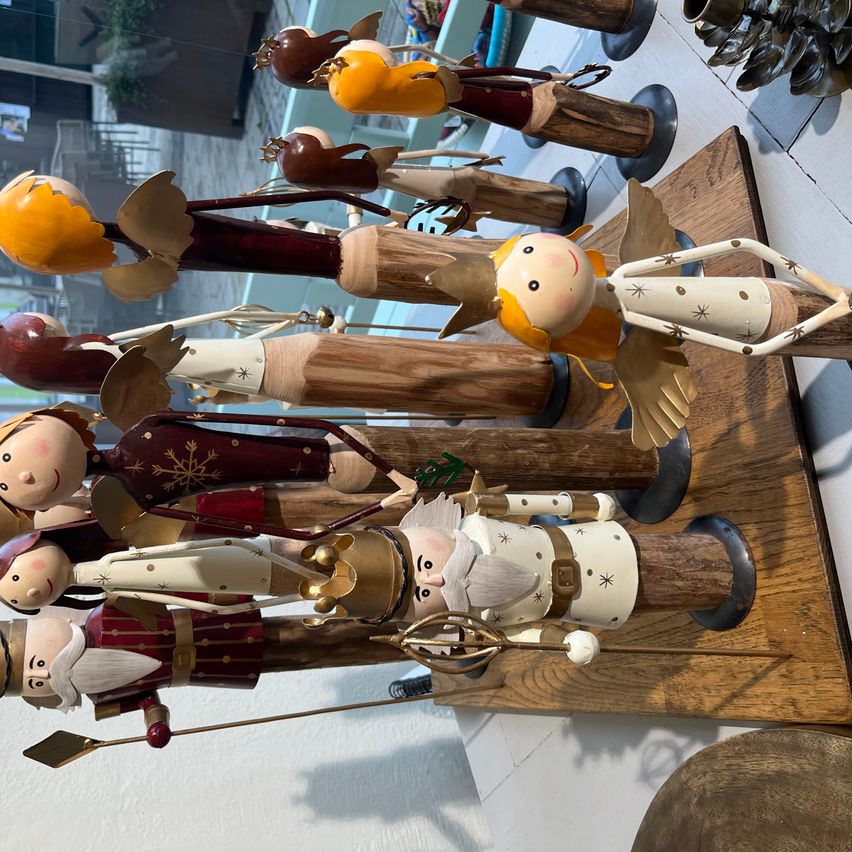 Ein Holzbrett zeigt verschiedene kunstvoll gestaltete Figuren. Die Figuren haben unterschiedliche Merkmale wie Flügel, Hüte und Schuhe. Einige sind in leuchtenden Farben bemalt, während andere ein traditionelleres Aussehen haben. Sie stehen aufrecht auf dem Brett, das an einer weißen Wand montiert ist.